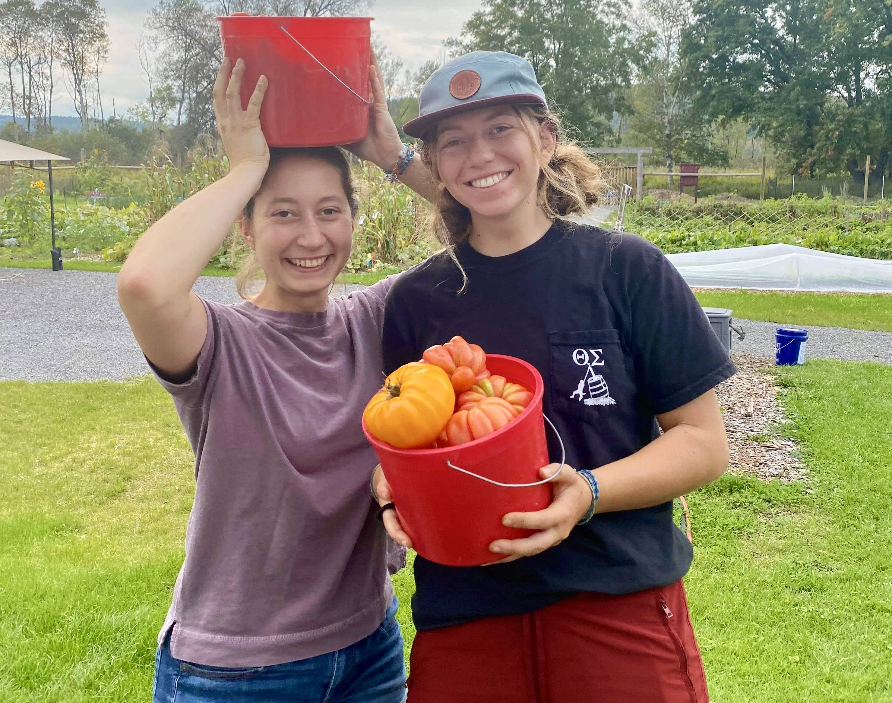 Two students in t-shirts show off their red buckets with freshly harvested pumpkins, tomatoes, and more.