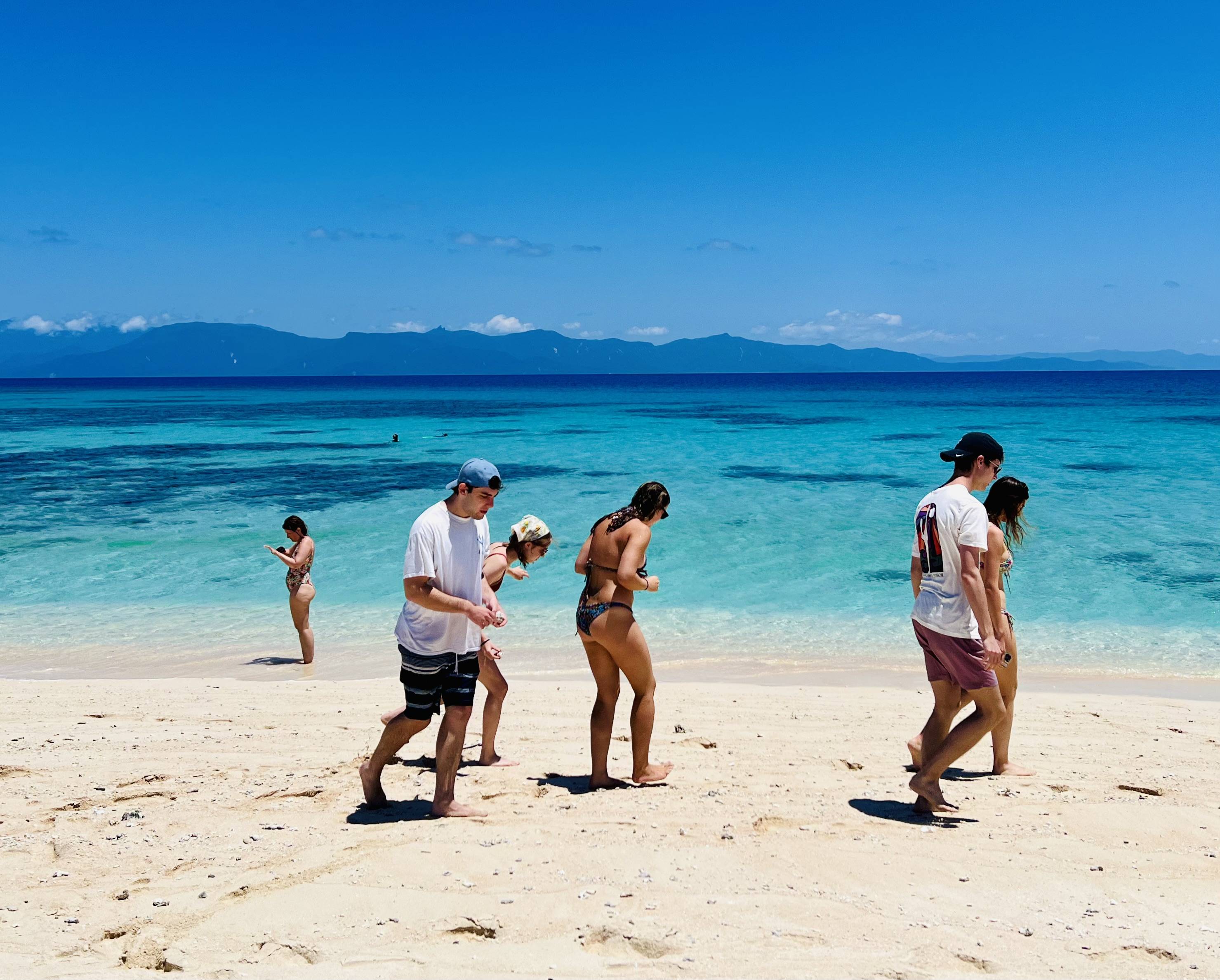 Students walking on a white sand beach with the bright blue ocean behind them.