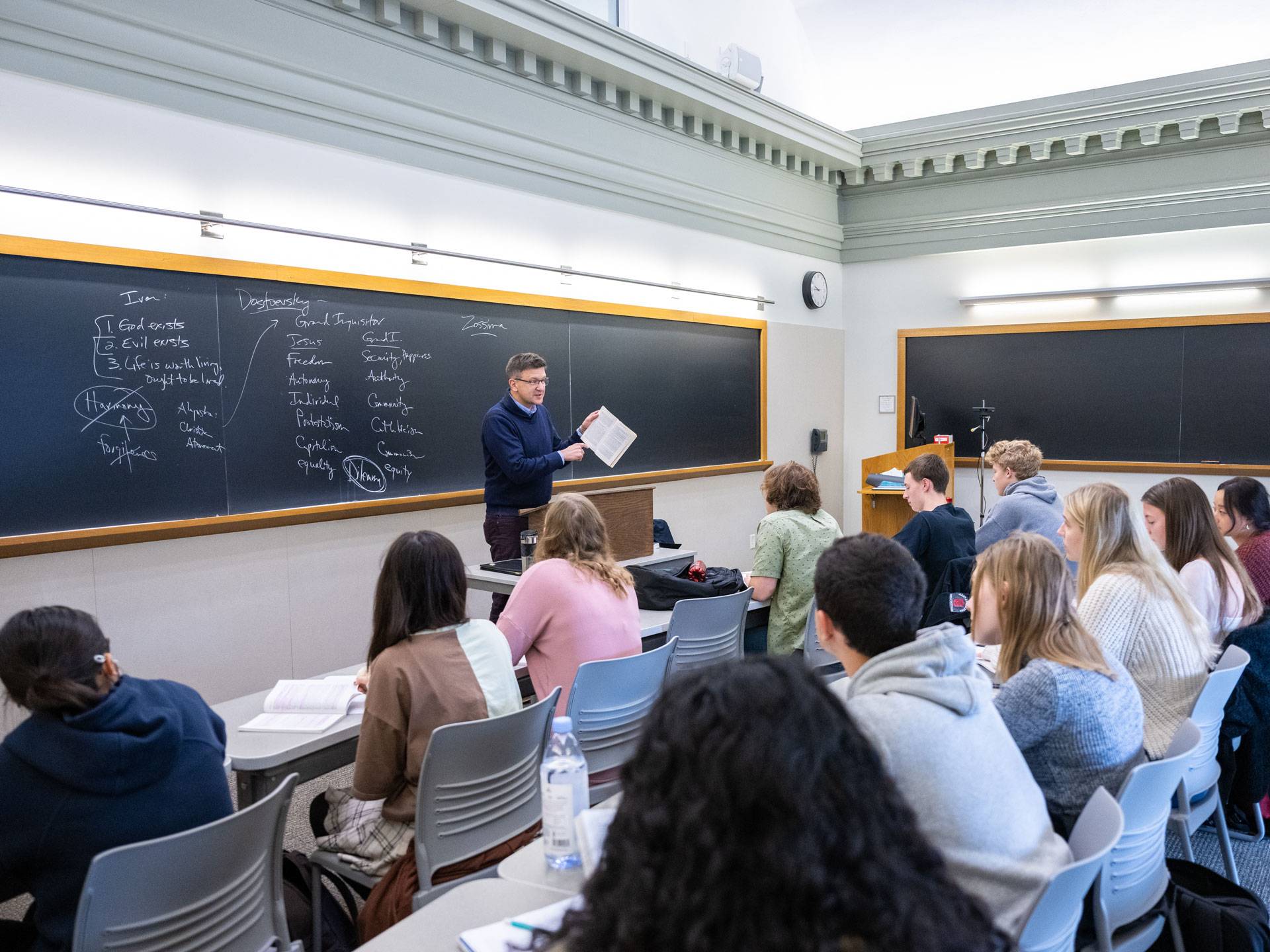 A male professor in a blue sweater in front of a blackboard instructs a class of students