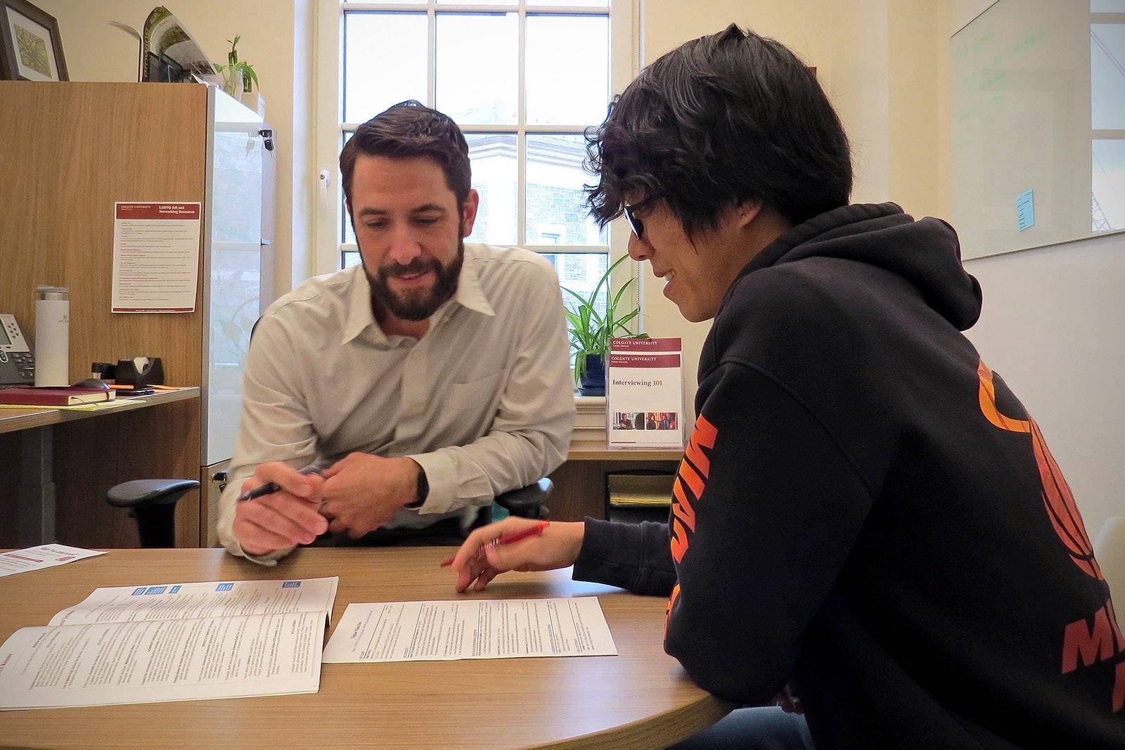 A career adviser sits at a desk with a student, going over some documents in a sunny room