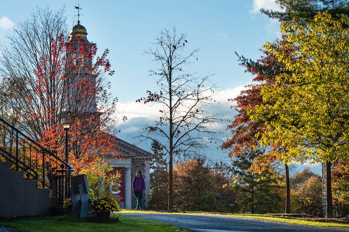 A person walks on a path in a park during autumn, with colorful trees and Colgate Memorial  Chapel in the background.